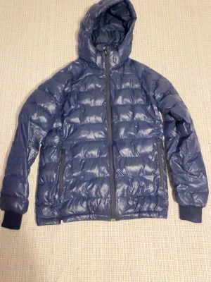 Mörkblå pufferjacka med huva - Snygg mörkblå pufferjacka med huva och dragkedja framtill. Jackan har två sidofickor med dragkedja och ribbade muddar vid ärmsluten. Materialet är glansigt och quiltat för extra värme. Perfekt för kalla dagar.