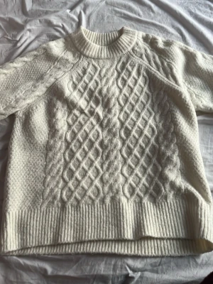Vit kabelstickad tröja - Säljer en vit kabelstickad tröja med klassiskt mönster och rund halsringning. Tröjan har ribbade muddar vid ärmslut och nederkant samt långärmad design. Materialet ser ut att vara ull eller ullblandning, perfekt för kyliga dagar.