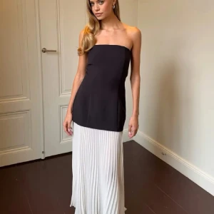 Svart och vit axelbandslös långklänning - Axelbandslös långklänning med svart överdel och vit plisserad nederdel. Klänningen har en stilren och elegant design med rak passform och ärmlös siluett. 