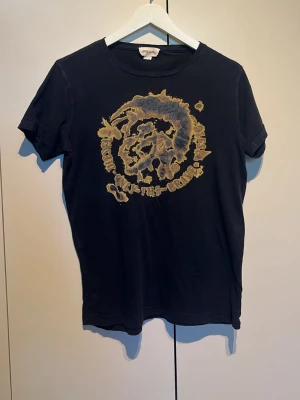 Svart t-shirt med tryck från Diesel - Cool svart t-shirt från Diesel med ett stort, unikt tryck i beige och grått på bröstet. Klassisk rund hals och korta ärmar. Tillverkad i mjuk bomull som känns skön mot huden. Perfekt för dig som gillar streetwear och vill sticka ut lite extra.