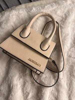 Beige Jacquemus Le Chiquito väska - Säljer en beige Jacquemus Le Chiquito handväska i ministorlek. Väskan har ett stilrent och strukturerat utseende med guldfärgad logga framtill, två handtag och en lång axelrem. Materialet är slätt och väskan har svarta kontrastsömmar som ger en cool touch.