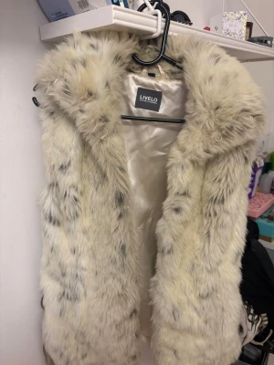 Beige fluffig pälsväst från Livelo - Supersnygg beige pälsväst från Livelo med mjukt, fluffigt fuskpälsmaterial och ljus, glansig insida. Västen har en öppen front och en cool, lite oversized look. Perfekt för att lyfta vilken outfit som helst med extra stilpoäng.