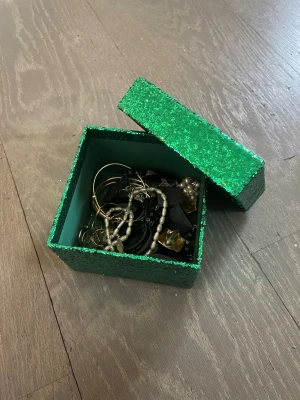 Smyckesmix i grön glittrig ask - Säljer en samling smycken i en grön glittrig ask. Innehåller armband, halsband och ringar i olika färger som guld, silver, svart och pärlvit. Blandade material som metall, plast och pärlor. Perfekt för dig som gillar att mixa och matcha accessoarer. Obs arbandet är äkta silver