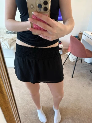 Svart kortkjol med smockad midja - Enkel och stilren svart kortkjol med bred smockad midja som ger en bekväm och flexibel passform. Kjolen har en flowy känsla och är gjord i ett lätt material, perfekt för varma dagar. Inga synliga mönster eller extra detaljer. Har sytt om den så den är kortare än originalet.