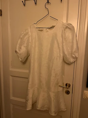 Vit kort klänning med puffärm - Supersöt vit kort klänning med puffärmar och volangkant nertill. Klänningen har öppen rygg med en stor rosett upptill och är tillverkad i ett glansigt tyg som ger en lyxig känsla. Perfekt för Studenten! Aldrig använd och har prislappen kvar.