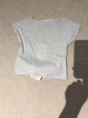 Ljusgrå topp med öppet rygg och knyt bak - Ljusgrå topp i mjuk bomull med korta ärmar och rund halsringning. Baksidan har öppet rygg med rosett längst ned på ryggen. Den kan vara lite som en kropptopp. Perfekt till jeans eller kjol för en enkel men snygg stil.