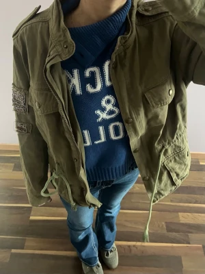 Olivgrön parkajacka med snörning - Snygg olivgrön parkajacka med snörning i midjan och axelklaffar. Jackan har dekorativa detaljer på ärmen och flera fickor framtill. Perfekt för dig som gillar en avslappnad och cool stil.