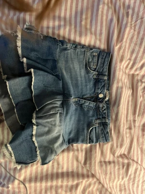 Blå jeansskort med volang och fransar - Säljer en blå jeansskort med volangdetaljer och råa fransar längs kanterna. Klassisk femficksmodell med knapp och dragkedja framtill. Skorten har en cool, avslappnad vibe och är gjord i denim. Perfekt för dig som vill ha något unikt och trendigt i garderoben!