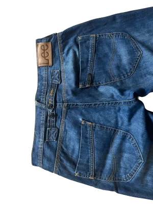 Blå raka jeans från Lee - Snygga blå jeans från Lee med klassisk femficksmodell och raka ben. Jeansen har slitningar framtill för en cool look och är gjorda i ett tåligt denimtyg. Perfekta för dig som gillar en avslappnad stil och vill ha ett par tidlösa jeans i garderoben. Jeansen passar 32/32 och har en riktigt unik fickdesign och justerbar midja