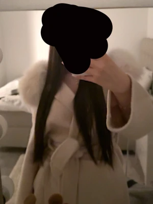 Beige kappa med äkta päls - Stilren beige kappa med stor päls runt kragen. Knytskärp i midjan ger en snygg siluett och kappan har en elegant, lång modell. Perfekt för dig som vill ha en trendig och mysig ytterjacka till hösten eller vintern. Köpt för 2300