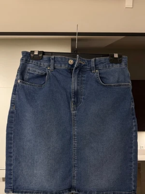 Blå jeanskjol med fickor - Säljer en klassisk blå jeanskjol med fem fickor och knappstängning fram. Kjolen har en rak passform och är tillverkad i denim.