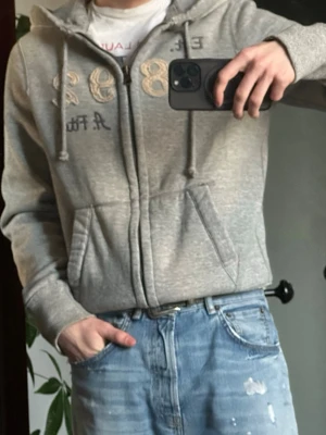 Grå hoodie från Abercrombie & Fitch - Snygg grå zip-hoodie från Abercrombie & Fitch med broderade detaljer och tryck framtill. Hoodien har dragsko i huvan, två fickor och ribbade muddar. Perfekt för en avslappnad och trendig look. Materialet är mjukt och skönt, troligen bomullsblandning.