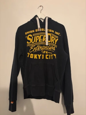 Mörkblå Superdry hoodie med tryck - Snygg mörkblå hoodie från Superdry med stort gult tryck på bröstet där det står 'Tokyo City'. Hoodien har vit dragsko i huvan och en klassisk känguruficka framtill. Tillverkad i mjukt bomullsmaterial och har ribbade muddar vid ärmar och nederkant.