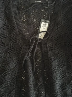 Svart stickad kofta från Vero Moda - Snygg svart stickad kofta från Vero Moda med knytning framtill. Koftan har ett genombrutet mönster och ribbade muddar vid ärmsluten. Perfekt att slänga över en topp för en chill look. Materialet är mjukt och följsamt.Koftan är dock lite liten i storleken och aldrig använd.