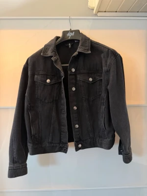 Svart jeansjacka med silverknappar - Snygg svart jeansjacka med klassisk krage och silverfärgade knappar framtill. Jackan har två bröstfickor med lock och knapp samt raka ärmar. Perfekt för dig som vill ha en stilren och tidlös look.
