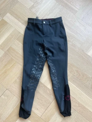 Mörkgrå ridbyxor med silikonprint - Snygga mörkgrå ridbyxor med helskodd silikonprint för extra grepp. Byxorna har bälteshällor, knapp och dragkedja framtill samt elastiska benslut med svarta och vinröda detaljer. Perfekta för dig som gillar stilrena och funktionella ridkläder.