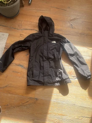 Svart regnjacka från The North Face - Svart regnjacka med huva från The North Face. Jackan har dragkedja framtill, två fickor med lock och justerbara ärmslut. Tillverkad i vattenavvisande material och har en stilren, sportig look med logga på bröstet.