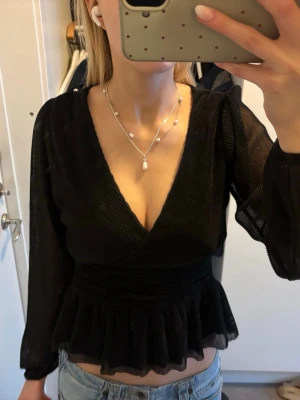 Svart omlottopp med volang - Snygg svart omlottopp med v-ringning och volang nertill. Toppen har långa, lätt transparenta ärmar och markerad midja. Perfekt att styla med jeans eller kjol för en trendig look.