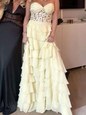 Ljusgul balklänning från zapaka  - Säljer en romantisk ljusgul långklänning med volanger i flera lager och spetsdetaljer upptill. Klänningen har hjärtformad byst och är ärmlös, perfekt för speciella tillfällen. Supersnygg och unik design som verkligen sticker ut. Aldrig använd