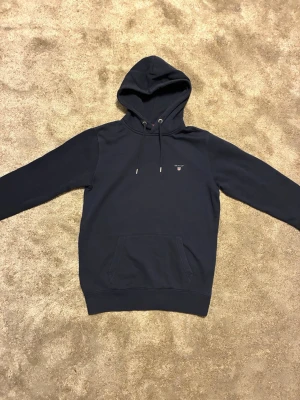 Mörkblå hoodie från Gant - Snygg mörkblå hoodie från Gant med liten logga broderad på bröstet. Klassisk känguruficka framtill och justerbar huva med dragsko. Tillverkad i mjuk bomullsmix som känns skön mot huden. Perfekt för en avslappnad och stilren look.