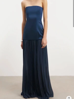 Mörkblå axelbandslös maxiklänning - Elegant mörkblå axelbandslös maxiklänning med figurnära överdel och plisserad kjol. Klänningen har en slät, glansig yta upptill och ett mjukt, flytande tyg nedtill som ger en lyxig känsla. Perfekt för dig som vill sticka ut med en stilren och modern look.