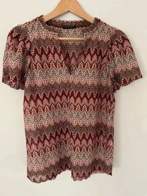 Mönstrad blus från Sisley - Säljer en snygg blus från Sisley med korta ärmar och ett färgstarkt mönster i vinrött, rosa, beige och brunt. Blusen har en lätt v-ringning och är gjord i ett tunt, luftigt material som känns skönt mot huden. Perfekt för dig som gillar boho-vibbar och unika prints.