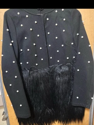 Svart kappa med pärlor och fuskpäls - Unik svart kappa dekorerad med vita pärlor över hela ovandelen och ärmarna. Nedre delen av kappan har en cool detalj i svart fuskpäls som ger extra edge. Rak passform och rund halsringning. Perfekt för dig som vill sticka ut.