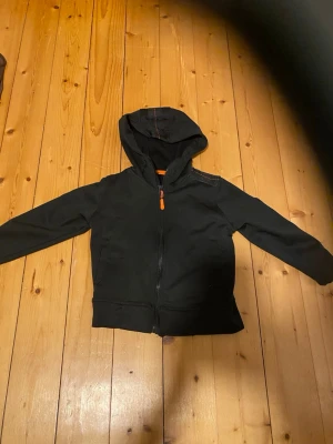 Svart hoodie med dragkedja och patch - Svart hoodie med hel dragkedja framtill och orange detaljer. Huva med dekorativ söm och en patch på bröstet med rymdtema. Mjuk och bekväm, perfekt för en avslappnad stil. Passar till jeans eller joggers.