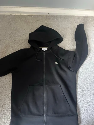 Lacoste hoodie. - Svart hoodie från Lacoste i klassisk passform med dragkedja framtill, två fickor och justerbar huva. Ikoniska Lacoste-loggan broderad på bröstet. Perfekt för en avslappnad och stilren look. Hör av er vid minsta fundering😁