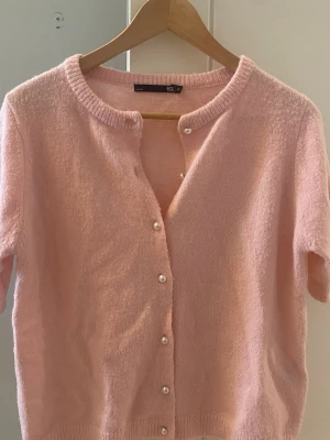 Rosa kofta med pärlknappar - Söt ljusrosa kofta från Lager 157 med korta ärmar och pärlknappar framtill. Koftan är stickad i mjukt material och har ribbade kanter vid hals, ärmslut och nederkant. Perfekt för en söt och enkel look. Priset kan diskuteras!🥰
