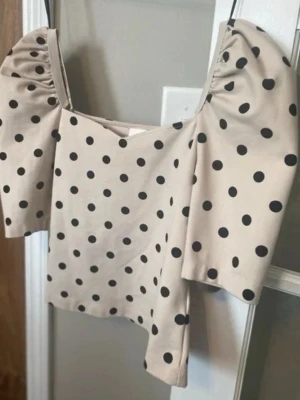Beige topp med puffärm och prickar - Trendig beige topp med svarta prickar och puffiga, korta ärmar. Fyrkantig urringning och figurnära passform ger en cool retrovibe. Perfekt att styla med jeans eller kjol för en snygg look.