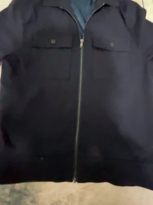 Mörkblå overshirt med dragkedja - Stilren mörkblå overshirt med dragkedja framtill och två bröstfickor med lock och knapp. Overshirten har en klassisk krage och raka ärmar. Perfekt att bära över en t-shirt för en avslappnad look.