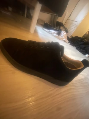 Svarta låga sneakers med röd sula - Säljer ett par svarta sneakers i mockaliknande material med lågt skaft och snygg röd sula undertill. Skorna har klassisk snörning och en stilren look som passar till det mesta. Perfekta för dig som gillar en clean och enkel stil.