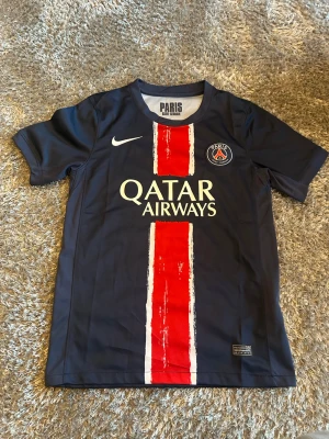 PSG matchtröja Nike mörkblå - Snygg mörkblå PSG matchtröja från Nike med röd och vit rand på framsidan, klubbmärke och Qatar Airways-tryck. Tillverkad i lätt och ventilerande material, perfekt för fotboll eller träning. Klassisk rund hals och korta ärmar. Har använts sparsamt 