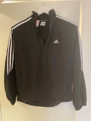 Svart vindjacka från Adidas - Svart vindjacka från Adidas med klassiska vita ränder längs ärmarna och logga på bröstet. Perfekt för sportiga dagar och streetstyle. Se bild för storlek.