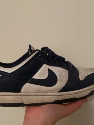 Nike Dunk Low blå och vita sneakers - Snygga Nike Dunk Low sneakers i blått och vitt läder med klassisk swoosh på sidan. Skorna har lågt skaft, vita paneler och mörkblå detaljer samt matchande blå snören. Perfekta för dig som gillar streetwear och vill ha en ikonisk look.