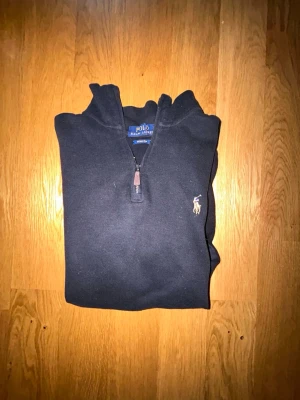 Svart halvzip-tröja från Polo Ralph Lauren - Svart långärmad tröja från Polo Ralph Lauren med halv dragkedja och klassisk broderad logga på bröstet. Tröjan har hög krage och ribbade muddar. Perfekt för en stilren och avslappnad look.