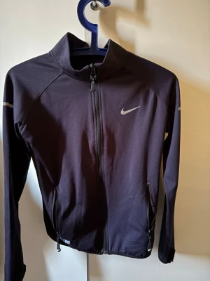 Svart träningsjacka från Nike - Snygg blå  träningsjacka från Nike med hel dragkedja framtill och två fickor med dragkedja. Jackan har en hög krage och diskreta reflexdetaljer på ärmarna samt Nike-logga på bröstet. Perfekt för löpning eller gym.