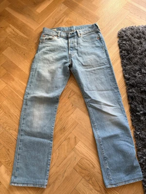 Levi's 501 ljusblå jeans - Klassiska Levi's 501 jeans i ljusblå denim med raka ben och femficksdesign. Jeansen har en snygg vintage-look med subtila slitningar och ikonisk läderpatch bak. Perfekt för dig som gillar tidlös stil och vill ha ett par jeans som alltid funkar.
