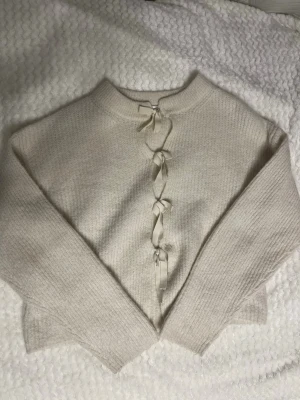 Stickad tröja med rosett - Snygg beige stickad tröja med långa ärmar och unika rosett-detaljer framtill. Tröjan har en rak, något kortare passform och ribbstickade muddar. Perfekt för dig som gillar en söt och trendig look.