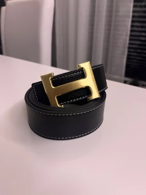 Svart skärp med guldfärgat H-spänne - Snyggt svart skärp i skinn med ett stort guldfärgat H-format spänne. Skärpet har vita sömmar längs kanterna som ger en stilren kontrast. Perfekt accessoar för att lyfta vilken outfit som helst.