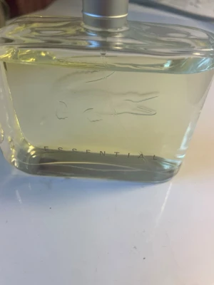 Lacoste Essential Eau de Toilette - Fräsch parfym från Lacoste i modellen Essential. Flaskan är rektangulär i genomskinligt glas med silverfärgad spraykork och den klassiska krokodilloggan ingraverad på framsidan. Doften är lätt gulaktig och flaskan har en stilren, modern design.