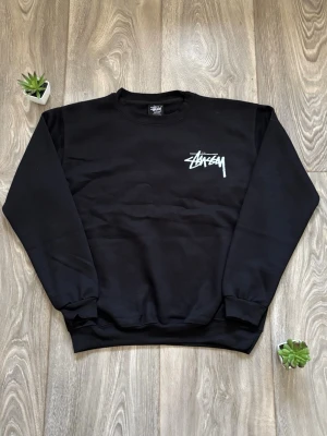 Svart Stussy sweatshirt - Svart sweatshirt från Stussy i perfekt skick med vit logga på bröstet och stort tryck på ryggen med jordglob, 8-ball och texten 'International Stussy Tribe'. Rund halsringning och ribbade muddar vid ärmslut och nederkant.