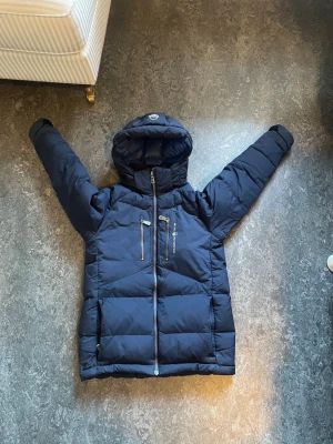 Marinblå pufferjacka från Sail Racing - Snygg mörkblå pufferjacka från Sail racing med huva och två snedställda bröstfickor med dragkedja. Jackan har en kraftig dragkedja framtill och är vadderad för extra värme. Perfekt för kalla vinterdagar och har en sportig look. Passar någon som är 140-155