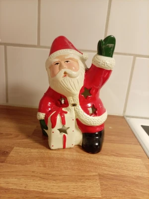 Keramik tomte ljuslykta - Säljer en keramik tomte ljuslykta med röd dräkt, vita detaljer och gröna vantar. Tomten har stjärnformade hål för ljusinsläpp och ett öppet fack på baksidan för att sätta in ett värmeljus. Perfekt som juldekoration. Den ä 18X12cm finns mer jul saker på min sida