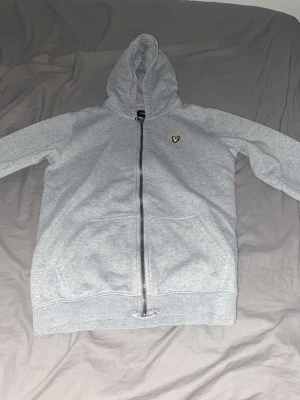Grå zip hoodie från Lyle & Scott - Ljusgrå hoodie med dragkedja från Lyle & Scott. Klassisk huva, två fickor framtill och den ikoniska gula örnloggan på bröstet. Tillverkad i mjukt sweatshirtmaterial som är perfekt för chill dagar. Enkel och stilren design som funkar till det mesta.