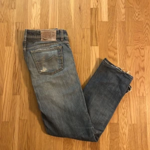 Blå slitna jeans från Jacob Cohen - Säljer ett par blå jeans från Jacob Cohen med slitningar och distressed detaljer. Jeansen har klassisk femficksmodell, knappgylf och en patch med logga bak i midjan. Materialet är denim i bomull med en snygg tvätt och casual look.