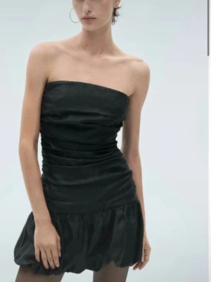 Svart axelbandslös kort klänning - Säljer en svart axelbandslös kort klänning med rynkade detaljer och volang nedtill. Klänningen har en tight passform upptill och är tillverkad i ett glansigt material som ger en elegant look. Perfekt för dig som vill ha en stilren och trendig outfit.