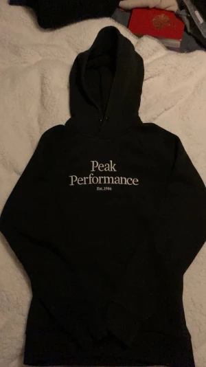 Svart hoodie Peak Performance S - Svart hoodie från Peak Performance med vit logga och text 'Est. 1986' på bröstet. Klassisk modell med huva och ribbade muddar. Perfekt för en avslappnad och sportig stil. Mjuk insida för extra komfort.