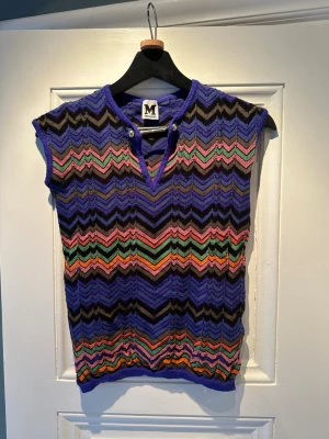 Mönstrad topp från Missoni - Färgglad topp från Missoni med ikoniskt sicksackmönster i lila, svart, rosa, grön, orange och brunt. Toppen är ärmlös med v-ringning och dekorativ säkerhetsnål vid halsen. Perfekt för dig som gillar unika och färgstarka plagg.  Storlek S/M (funkar då den är i stretch)  Bra skick.  En riktigt fin designerpiece som ger en väldigt cool vintage-look.  Observera: Jag har tyvärr inte kvar kvittot, men märkeslapp finns tydligt i plagget (se bild).